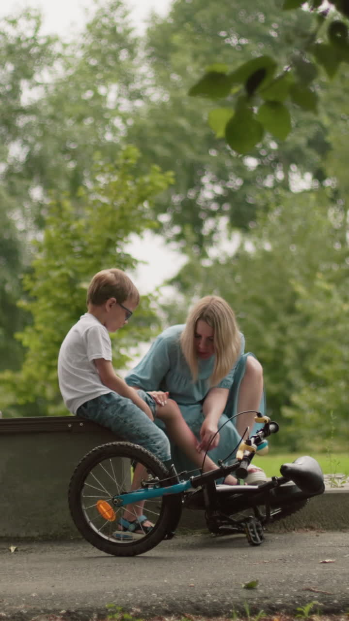una madre atiende suavemente la herida de su hijo mientras está sentada a lo largo de un camino asfalto en un parque, el niño, sosteniendo sus pantalones, parece triste mientras su madre le limpia la pierna