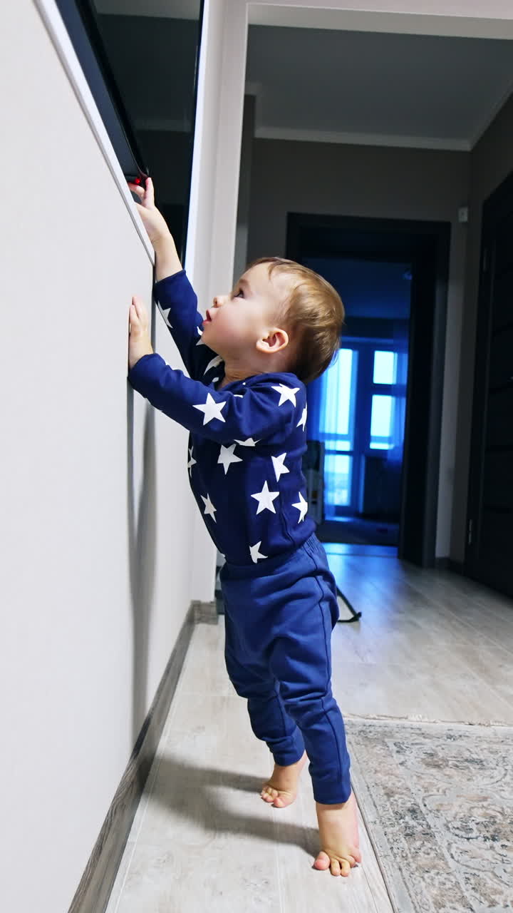 Caucasian toddler boy stands on his toes trying to reach the TV on the wall. Baby tries to switch on TV. Vertical video