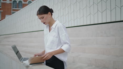 Girl-freelancer-working-laptop-outdoors-checking-information-in-papers-vertical