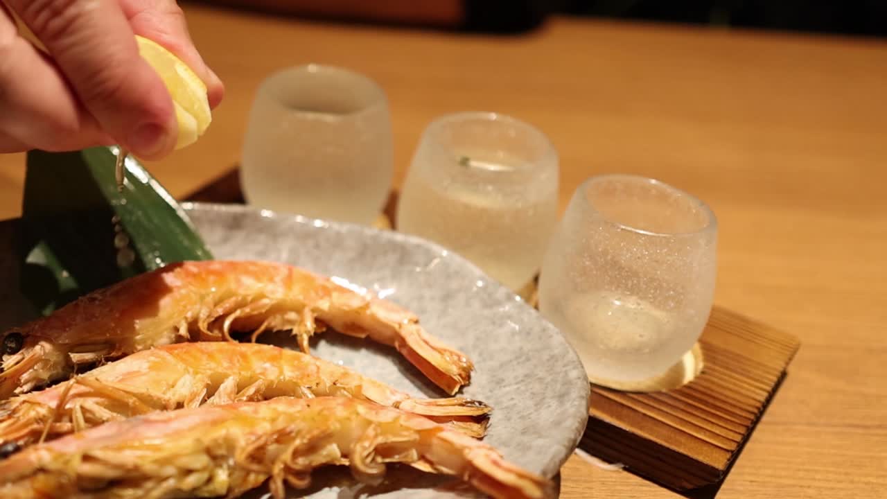 A hand squeezes lemon over grilled shrimp on a plate, with glasses in the background.