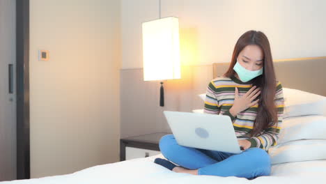 Asian-woman-on-self-isolation-quarantine-in-the-hotel-feel-bad-and-coughing-while-using-her-laptop-computer,-she-is-wearing-a-protective-surgical-mask