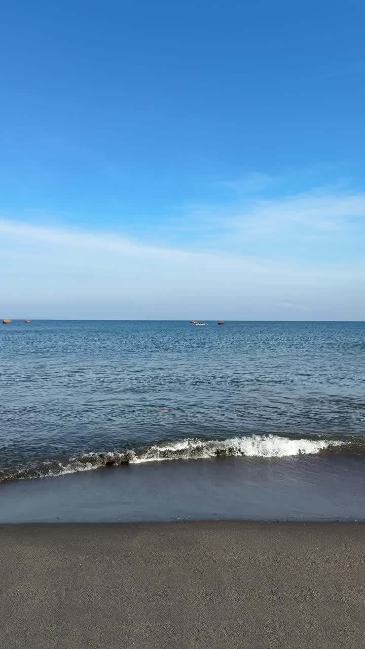 Ombak Laut yang Tenang di Pantai Pasir Gelap dengan Langit Biru.
