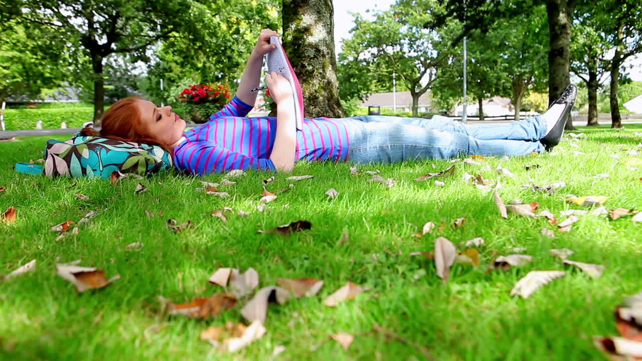 잔디 위에 누워 있는 예쁜 젊은 학생이 글을 쓰고 있다.