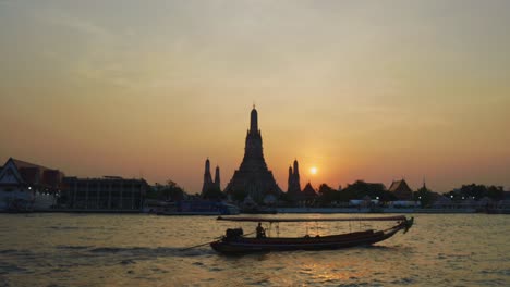 Wat-Arun,-temple-of-Dawn,-in-Bangkok-Thailand-with-a-Longtail-River-Boat-floating-by-at-Sunset