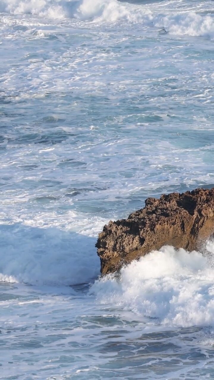 Potenti onde oceaniche si infrangono contro una roccia marrone solitaria in una composizione verticale.