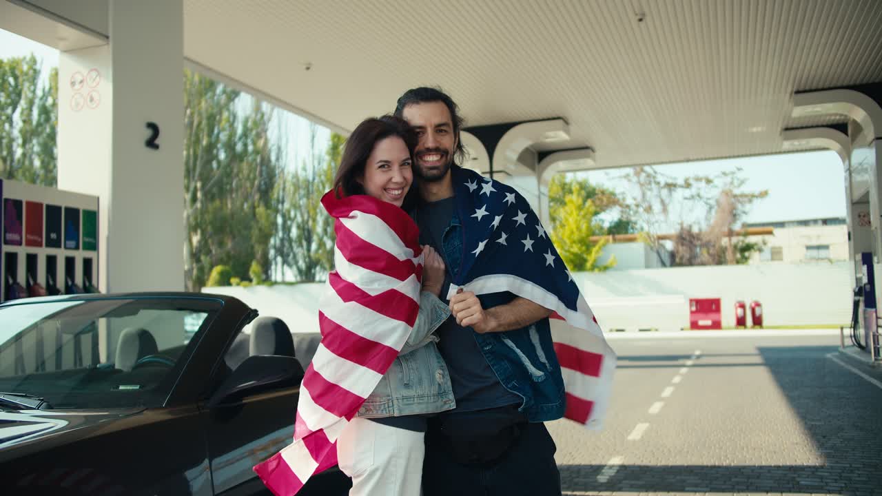 feliz chica morena besando a su novio, pareja envuelta en la bandera de los estados unidos y de pie en la gasolinera cerca de su descapotable gris
