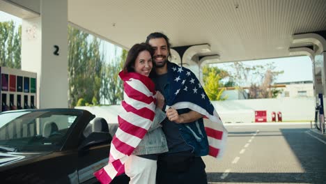 Happy-brunette-girl-kissing-her-boyfriend,-couple-wrapped-in-US-flag-and-standing-at-gas-station-near-their-gray-convertible