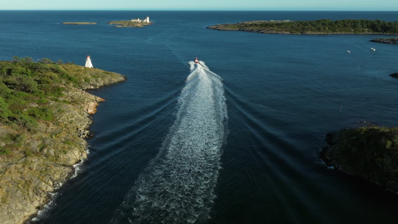 A vibrant red boat speeds across the serene blue water under a clear sky. A small island with a lighthouse is visible in the background, adding charm to the scene