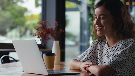 Young-Businesswoman-With-Takeaway-Coffee-Making-Video-Call-On-Laptop-In-Coffee-Shop