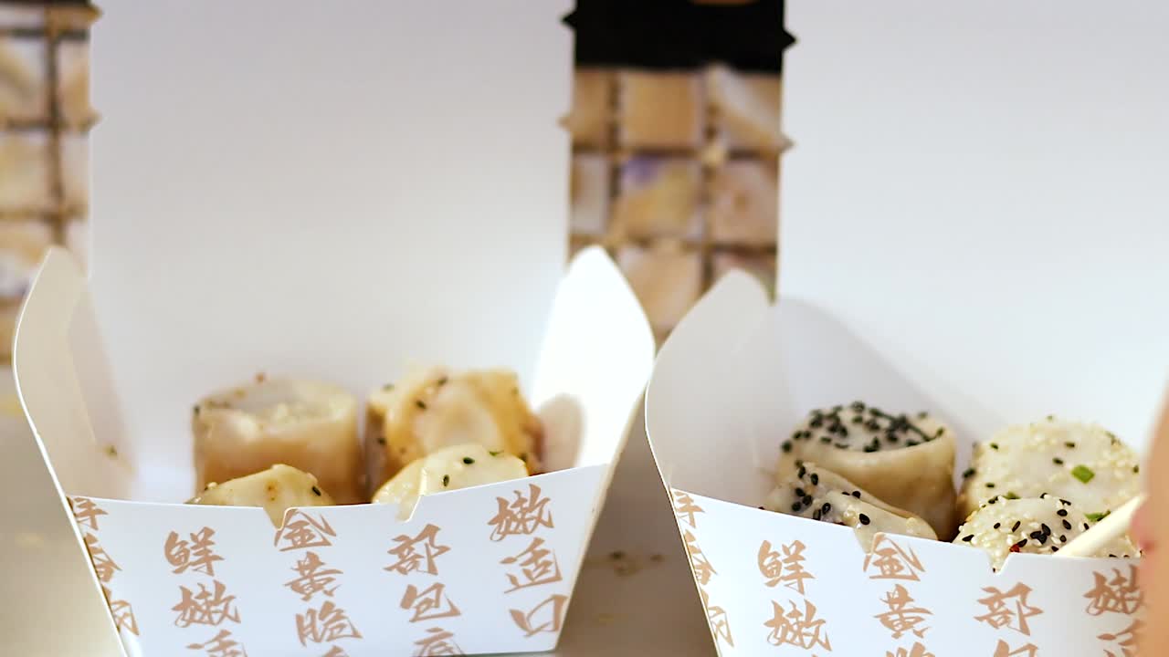 A close-up of chopsticks selecting sesame-topped buns from a branded takeout box.