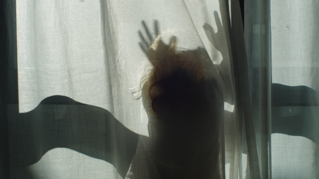 Shadowy female figure creating dramatic silhouette while waving behind translucent curtain, casting intricate shadows with soft sunlight streaming through delicate fabric