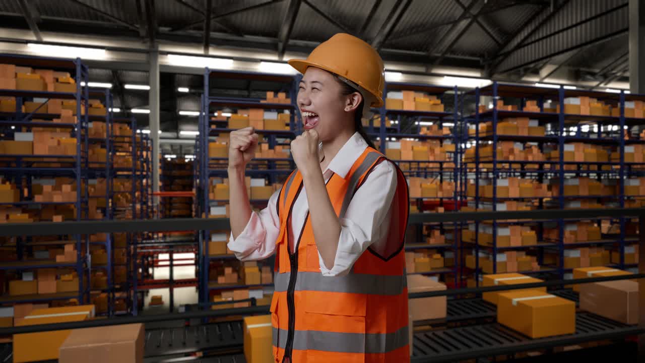 안전 헬을 입은 아시아 여성 엔지니어의 측면 뷰. 서서 비명을 지르는 골. 배달 상품으로 가득 찬 선반으로 창고에서 일하는 것을 축하하는.