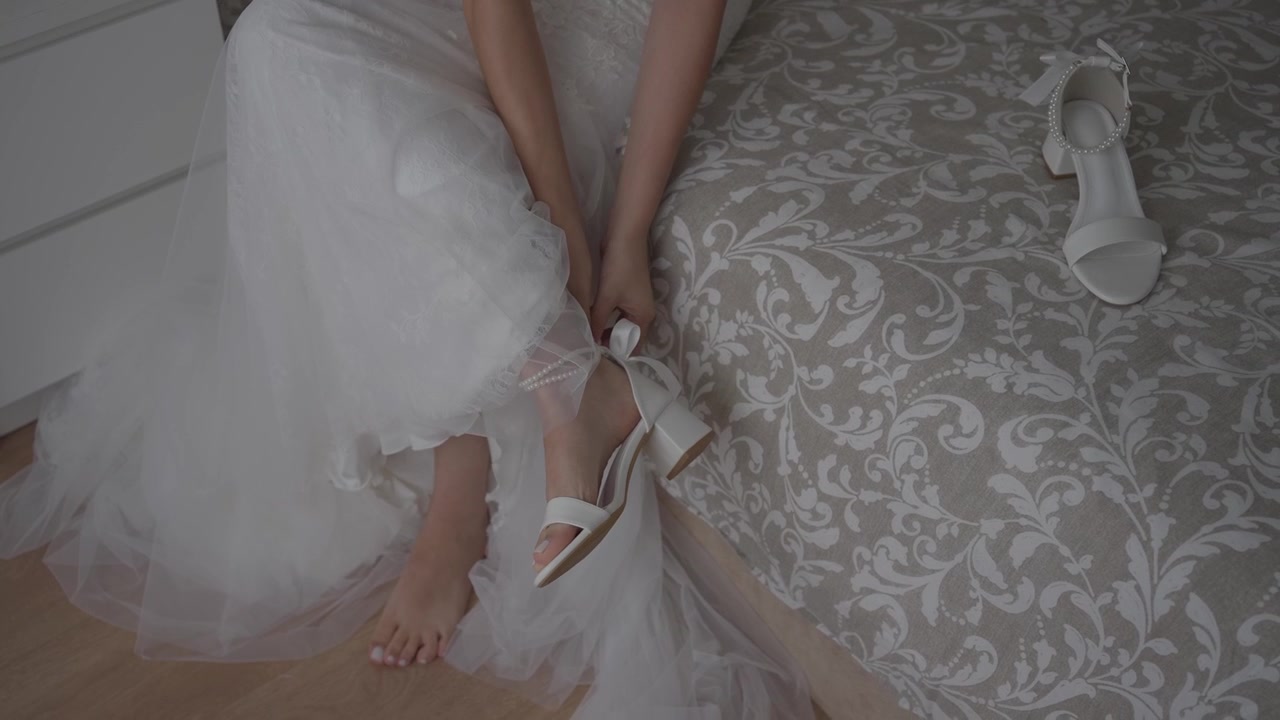Close view of bride wearing white heels while sitting on bed in dress