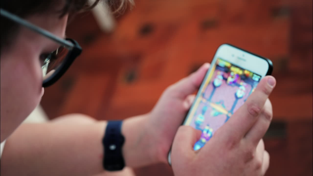 Over-the-shoulder close-up of a kid with glasses playing on a smartphone indoors