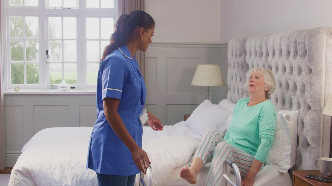 Senior Woman At Home Using Walking Frame Being Helped Out Of Bed By Female Care Worker In Uniform