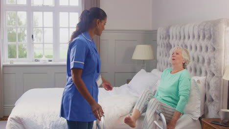 senior woman at home using walking frame being helped out of bed by female care worker in uniform