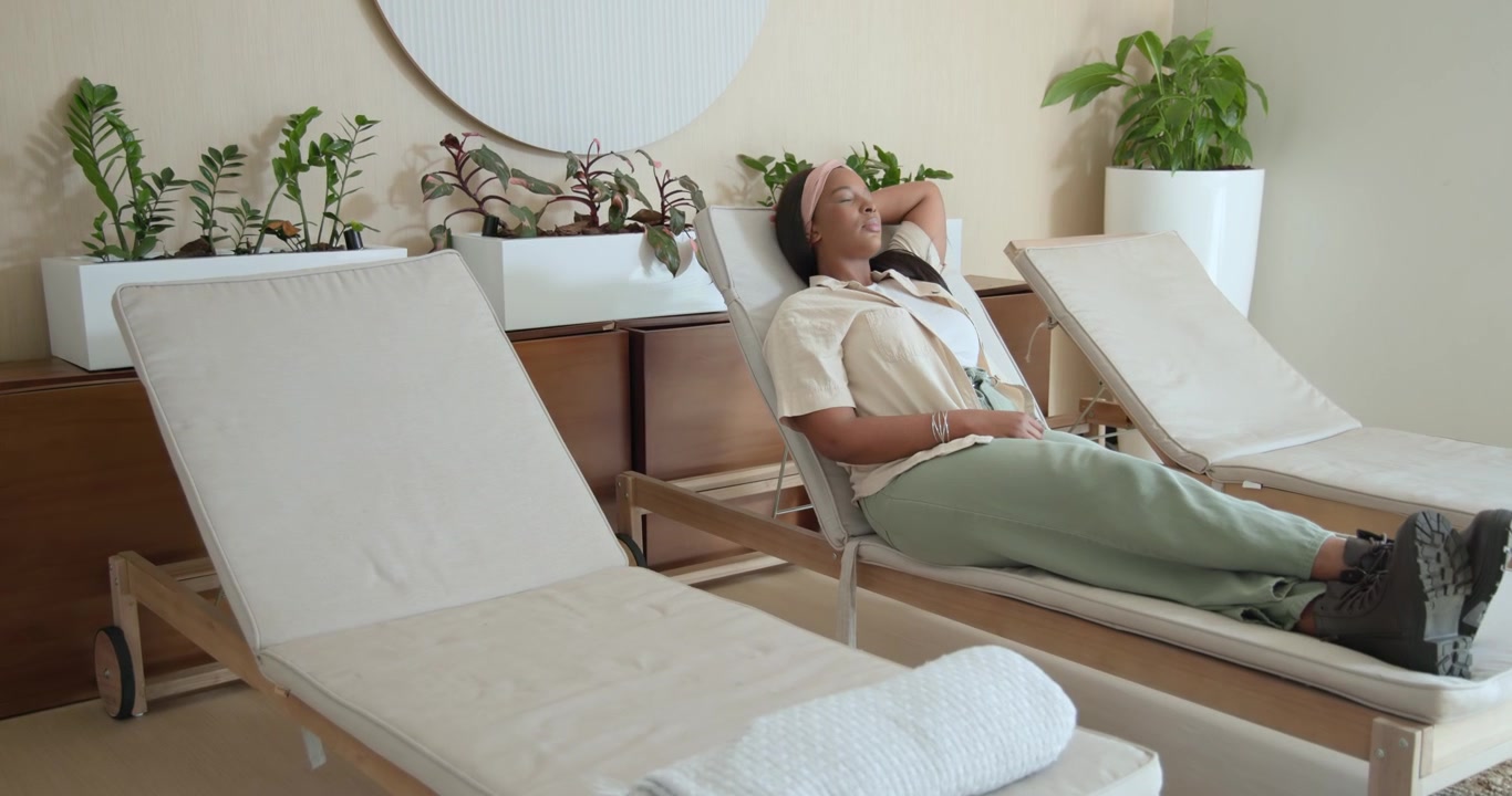 Donna afroamericana che entra in una sala spa, cammina verso una sedia, si siede, si sistema la fascia per capelli in cerca di riposo.