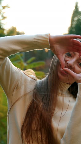 happy beautiful woman in white warm sweater outdoors. lady smiling to the camera and showing heart sign to the camera. autumn nature at backdrop.