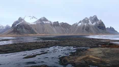 Volcanic-black-sand-beach-at-low-tide,-snowy-mountain-range-beyond