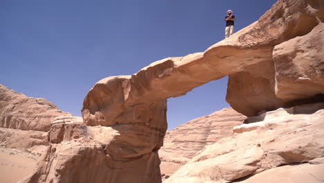 male standing on natural arch in desert landscape of wadi rum, jordan on hot sunny day, panorama