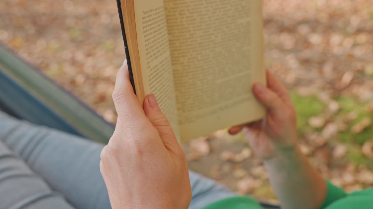 Lettore in campeggio con amaca che tiene un libro all'aperto, primo piano delle mani che girano le pagine mentre si rilassa in amaca al campeggio, luce solare filtrata sulla lettiera di foglie, gambe in denim e manica di camicia verde visibili, ritmo lento.