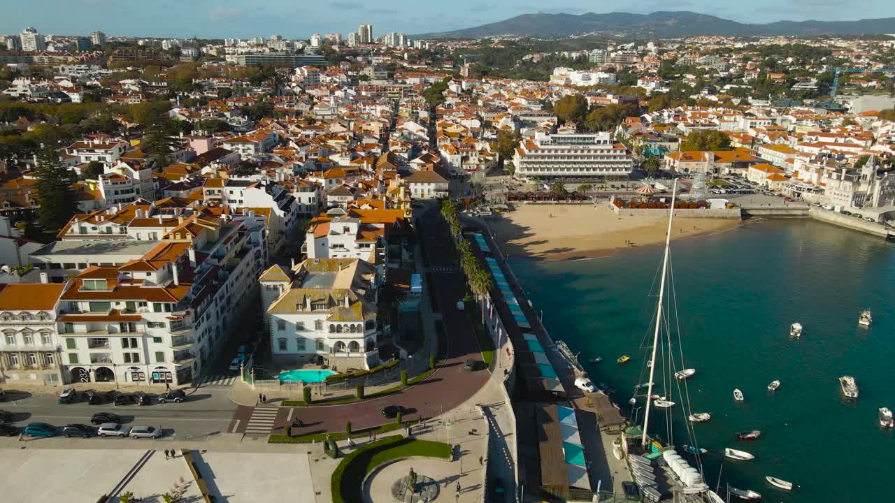 Helle Luftaufnahme von Cascais, die Boote, die Uferpromenade und den Sandstrand neben einer malerischen Küstenstadt an einem klaren, sonnigen Tag zeigt