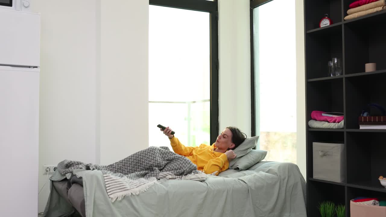 Giovane donna cambia canali TV con il telecomando mentre è sdraiata sul letto