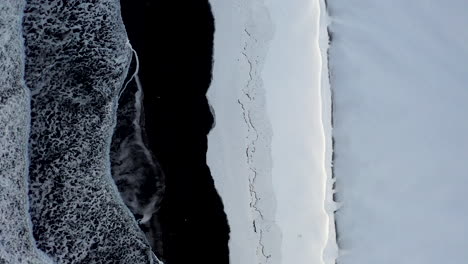 AERIAL:-Birdsview-of-Black-Beach-with-white-arctic-snow-in-Iceland-in-Winter-Snow,-Ice,-Waves,-Water