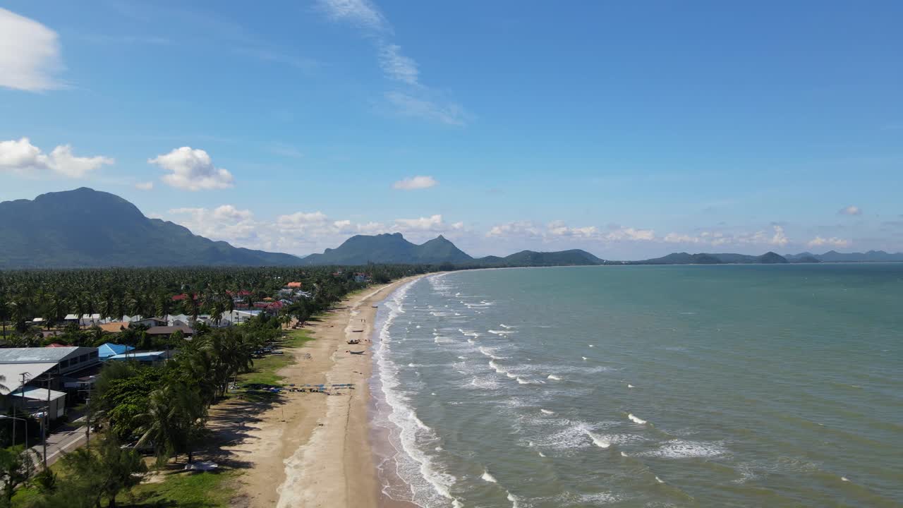 Metraje aéreo en retroceso a gran altitud de montañas en el horizonte, olas rompiendo, una carretera y cocoteros, frente a la playa