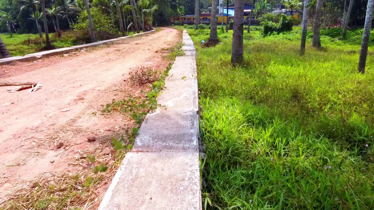 A serene tracking shot along a rural concrete boundary wall, bordering a red dirt path and a lush coconut grove, as a yellow train passes in the distance through tropical greenery