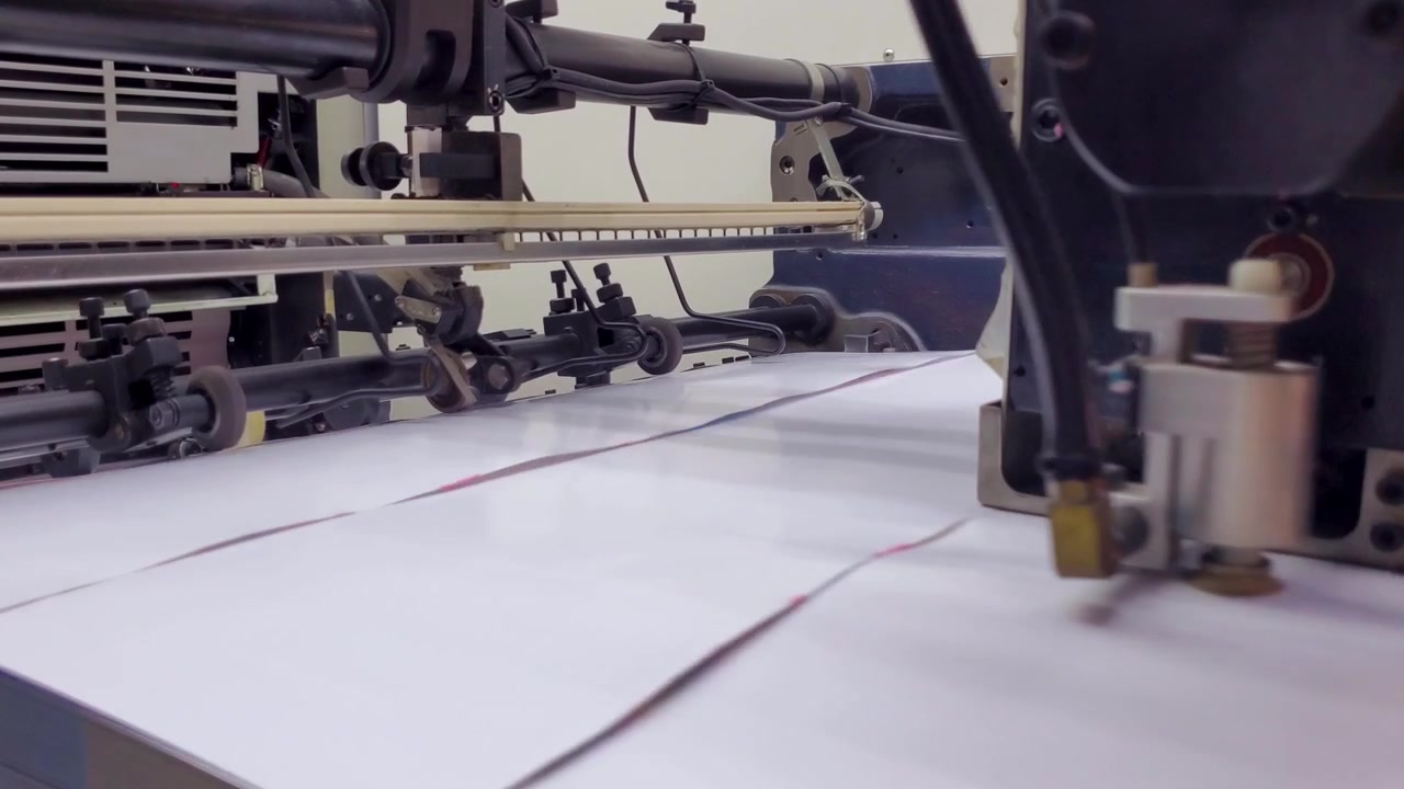 Close-up of a mechanical suction arm picking up individual white paper sheets in a high-precision printing press during a manufacturing process.
