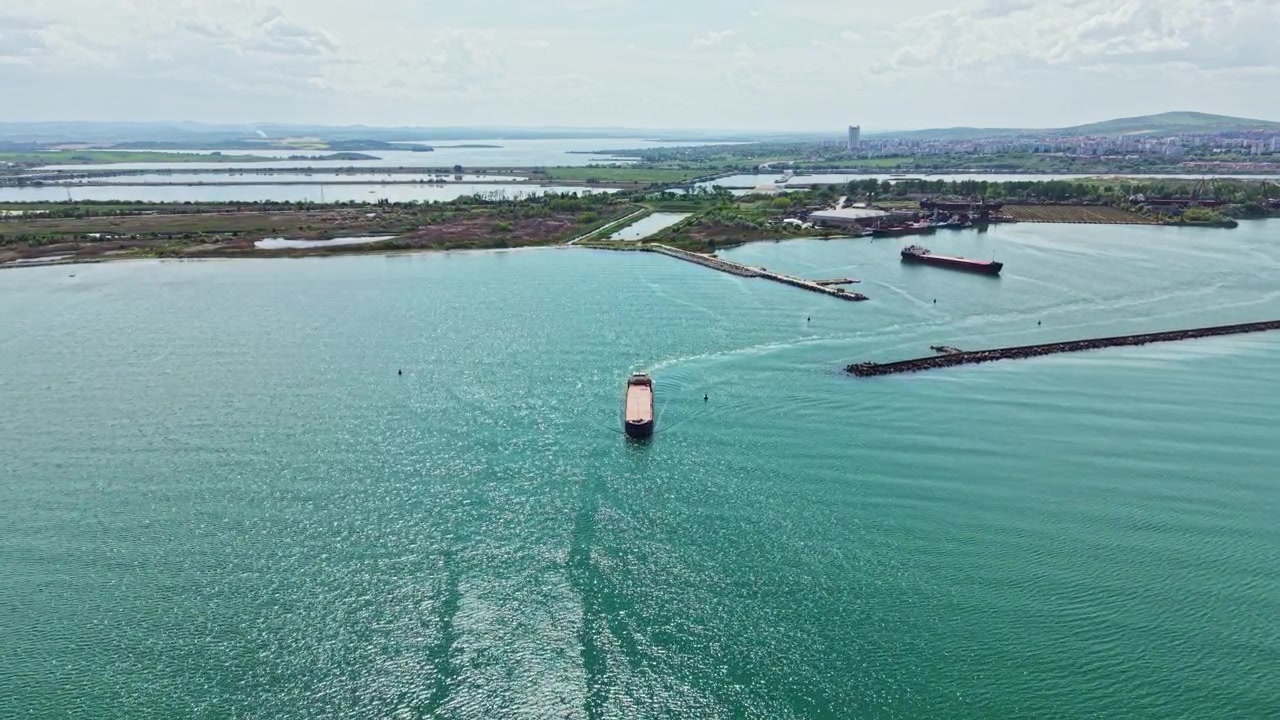 Vista di una grande nave da carico che naviga in acque blu vicino a una zona costiera con terra distante e strutture sullo sfondo durante il giorno.