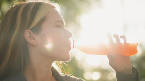 woman seated outdoors drinking orange juice with a refreshing expression, enjoying a moment of relaxation, background features soft glowing light and greenery