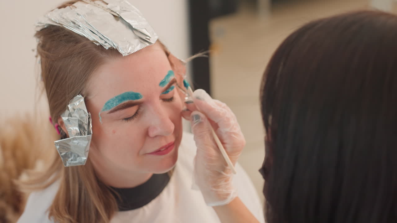 salon eyebrow tinting with turquoise pigment, stylist wearing gloves applies pigment to brows while hair wrapped in foil, closeup of client smiling during color process, professional tools visible