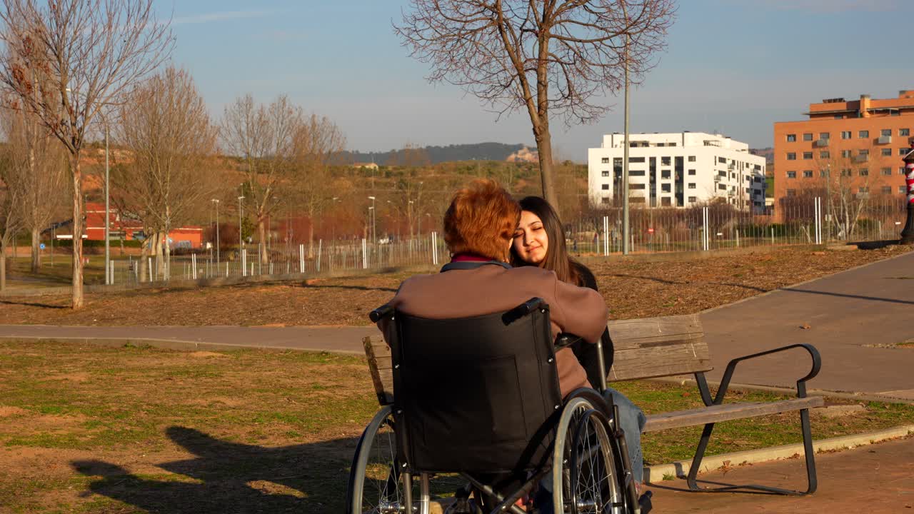 Rear shot of an elderly woman in wheelchair talking with granddaughter on park bench on sunny day