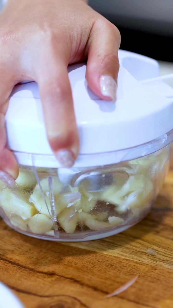 A hand operates a manual food processor to chop garlic on a wooden surface under bright lighting