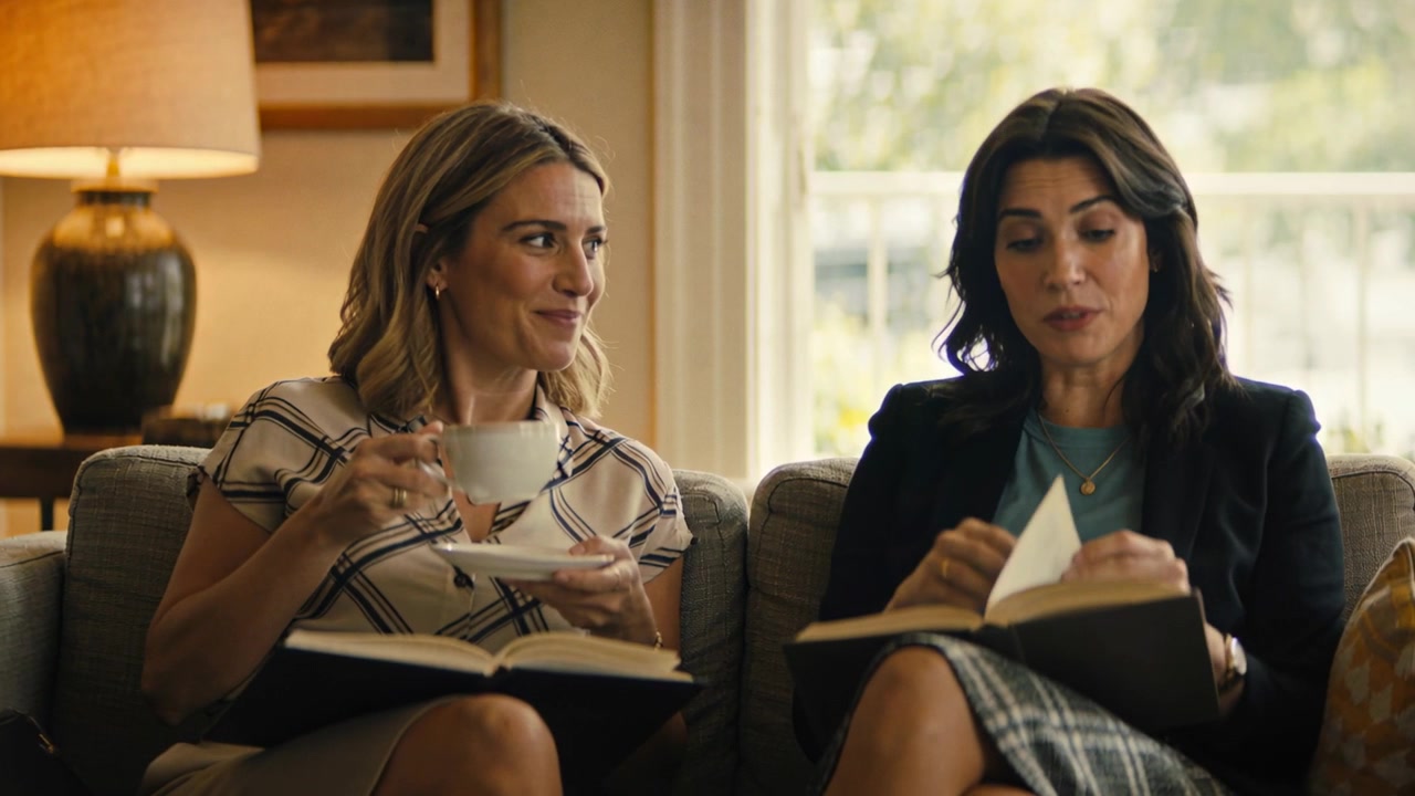 Lifting teacup, two women in office clothes discussing book, gesturing turning pages in living room