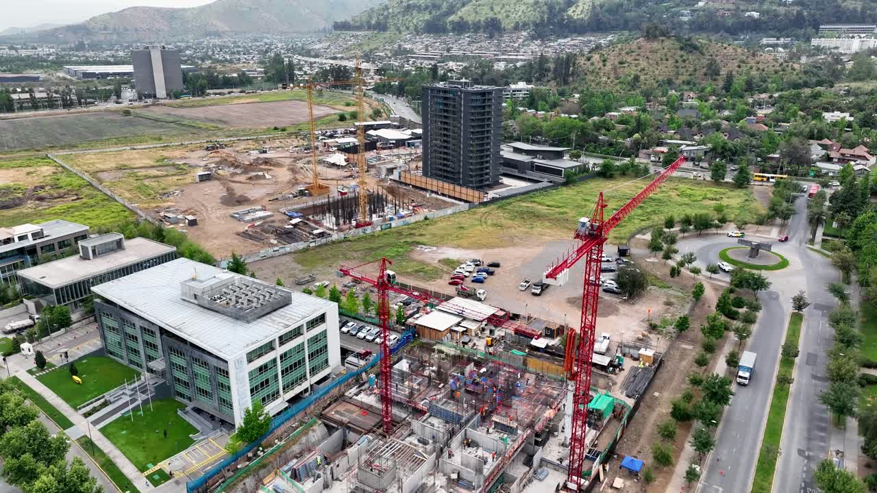 Forward drone aerial of construction site with red cranes, foundation work, high-rise tower, mountains backdrop, and urban landscape