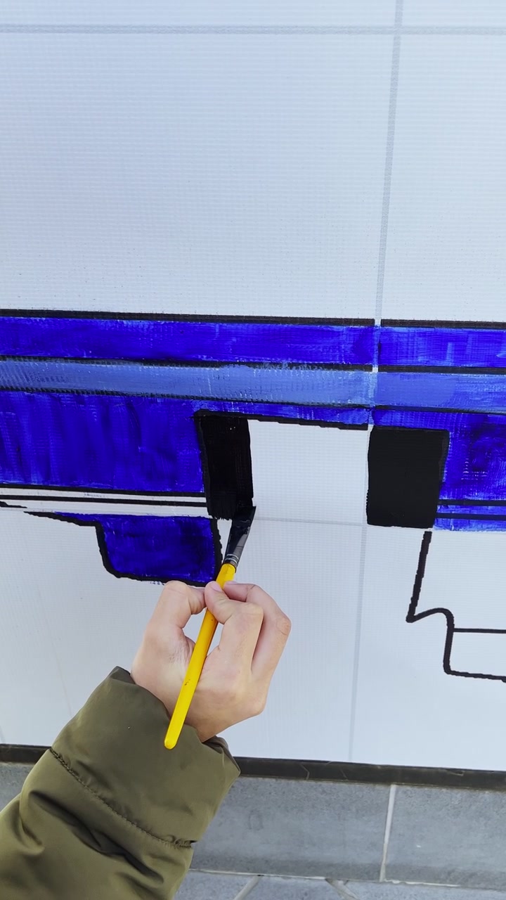 Close-up vertical da mão de uma pessoa usando um pincel amarelo para pintar formas geométricas pretas e azuis em uma parede de azulejos brancos. Criação de arte de rua ao ar livre durante o dia claro.