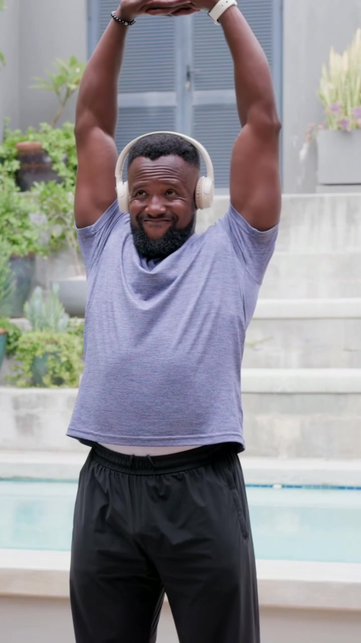 Vertical video: Starting man in purple tee doing warmup routine on patio, headphones, smartwatch