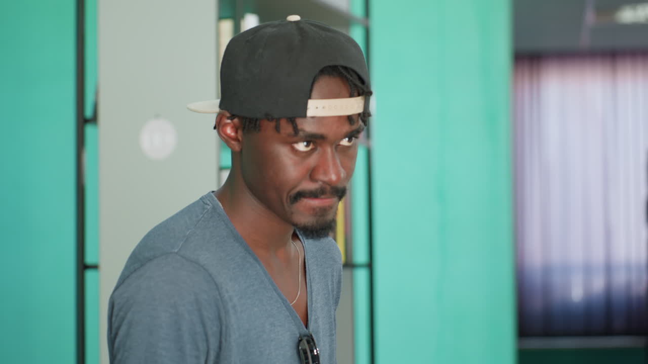 Young man in casual clothing wearing cap walks through library aisle, glancing thoughtfully at books on shelf against colorful background