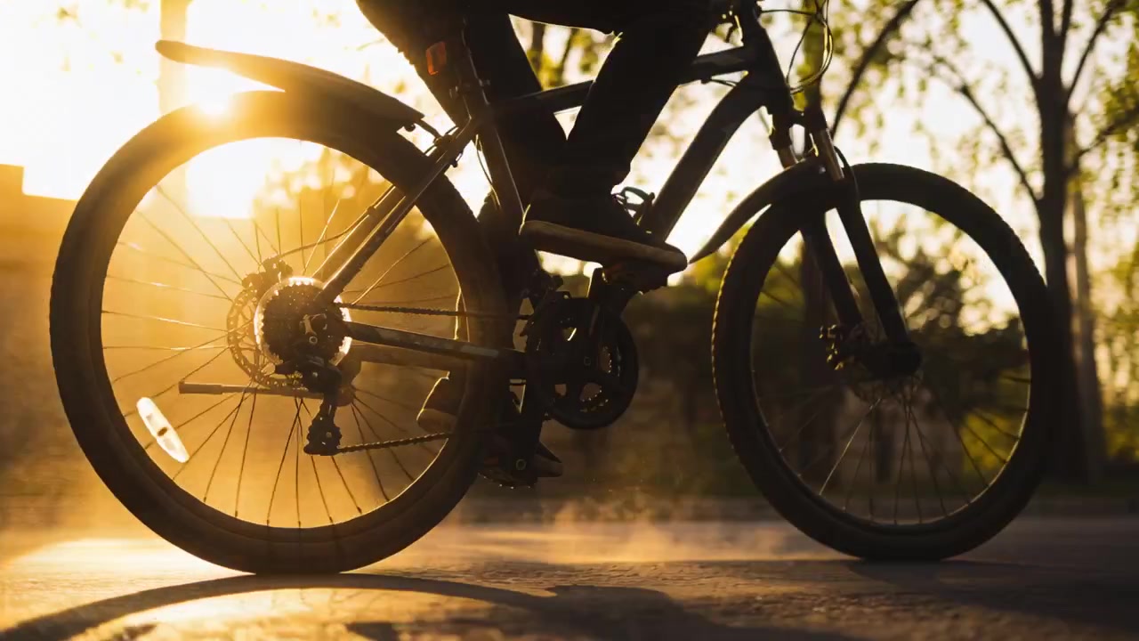 Cyklende cyklist i mørke bukser kører ad asfalteret vej, mens lav sol bagbelyser kæden, og støv hvirvler op.