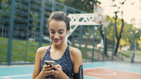 chica deportiva con airpods riéndose mientras envía un mensaje de texto en un teléfono inteligente en la cancha al aire libre en un día de verano