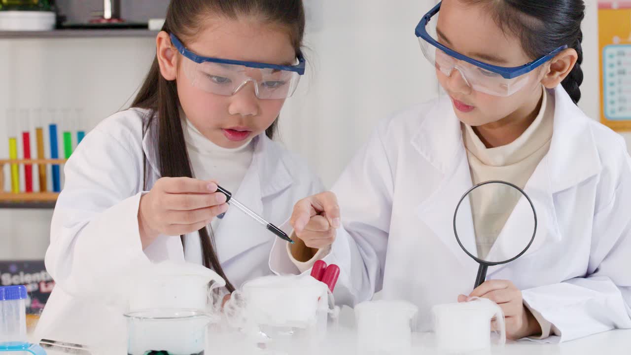 Dua gadis muda mengenakan jas lab dan kacamata pengaman saat melakukan percobaan kimia berasap