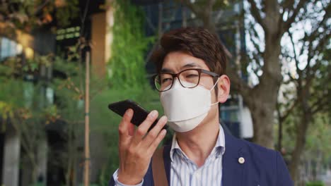 hombre asiático con máscara facial hablando en un teléfono inteligente mientras camina por la calle
