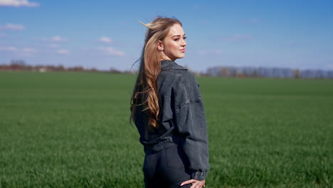 young woman outdoors. attractive girl in denim suit on green field. beautiful girl with long hair gets back on camera and smiles on a meadow. rear view.