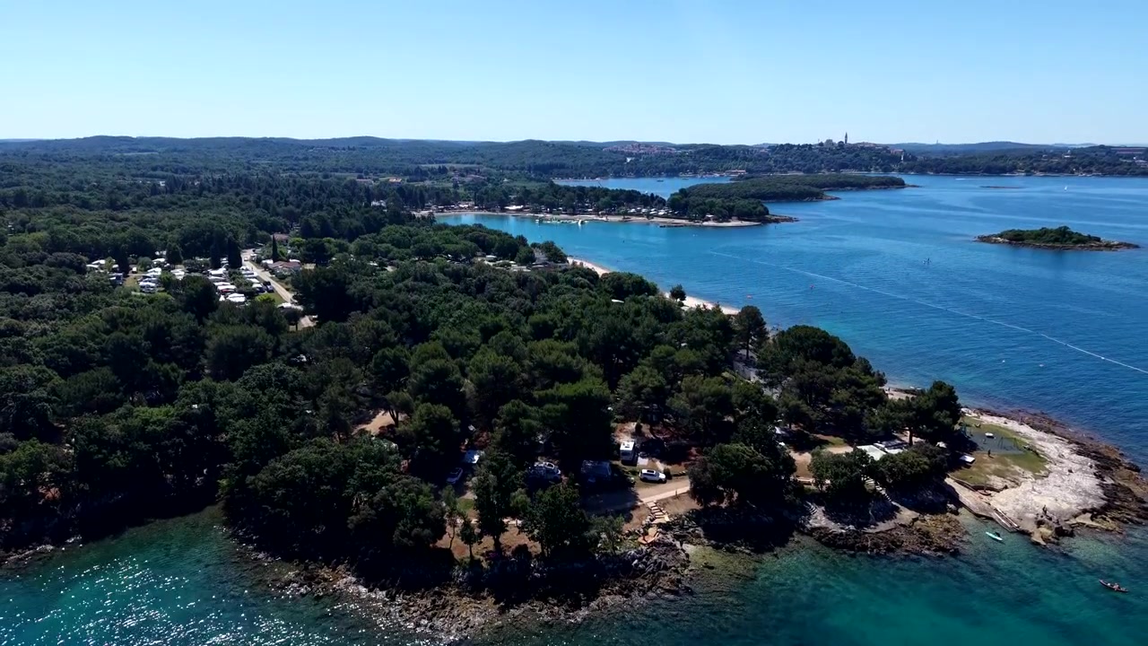Campeggio su penisola boscosa ospita roulotte immerse tra i pini, limpide acque blu dell'Adriatico e piccole isole che si estendono verso la lontana costa rocciosa sottostante. Croazia. Ampia ripresa aerea con drone in salita.
