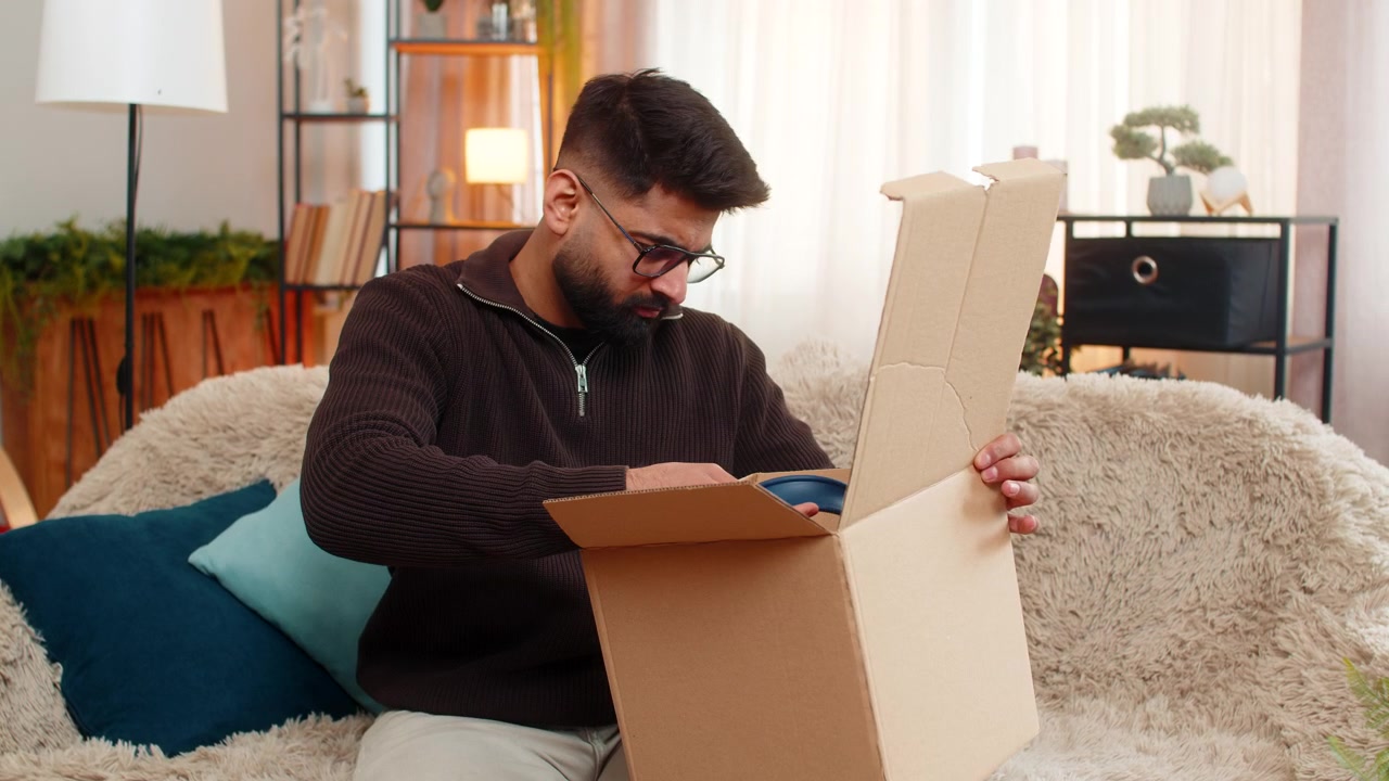 Indian man on sofa opens box and shakes head after discovering wrong item in online delivery order