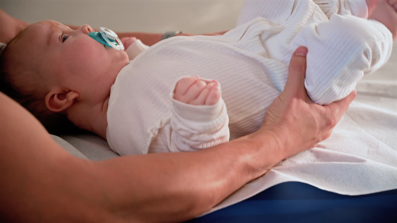 Primo piano di un neonato disteso tranquillamente su un lettino medico, che tiene in mano un ciuccio mentre un adulto lo sostiene delicatamente