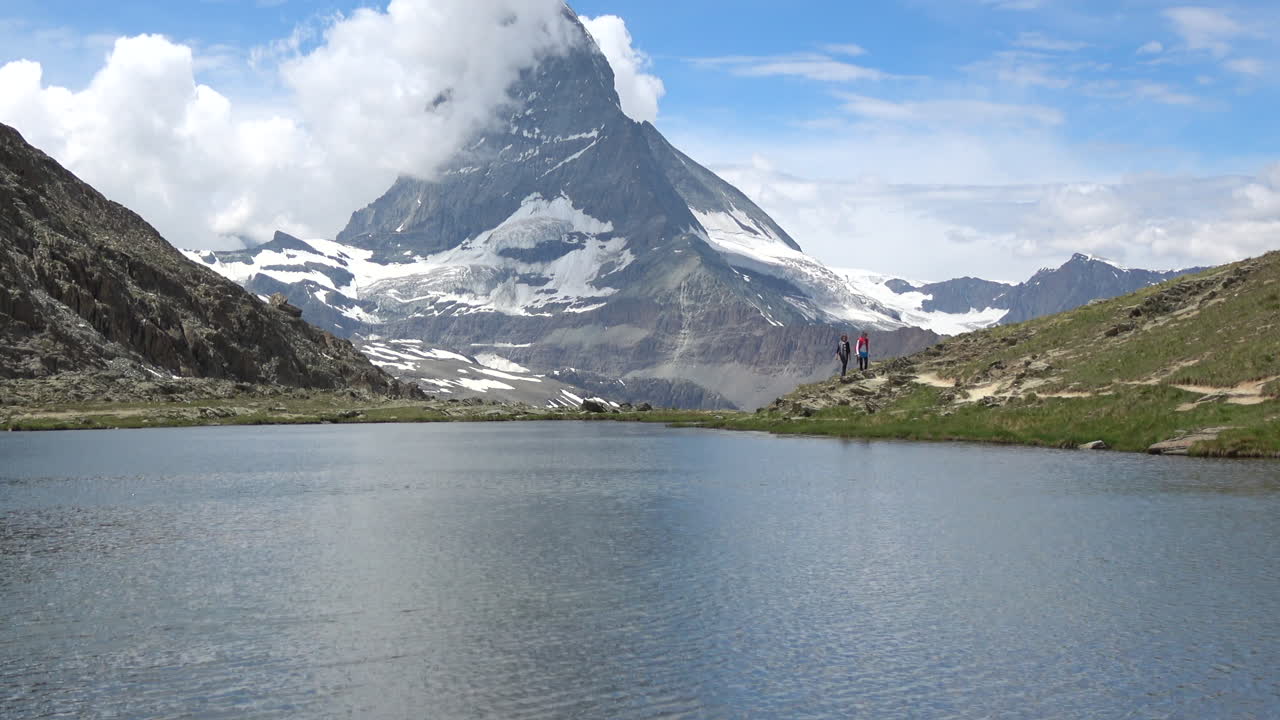 Ampia veduta di un lago alpino calmo che riflette il Cervino, con escursionisti sulla cresta, pendii verdi e nevai sotto le nuvole a Zermatt, Vallese, Svizzera.